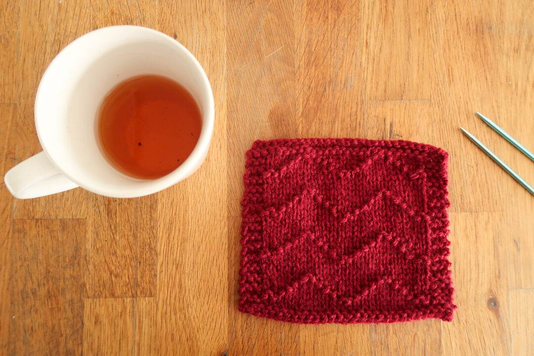 Wave Stitch knitting stitch for beginners. This red textured easy knit stitch features a chevron pattern, and is laying on a wooden table next to a mug of tea.