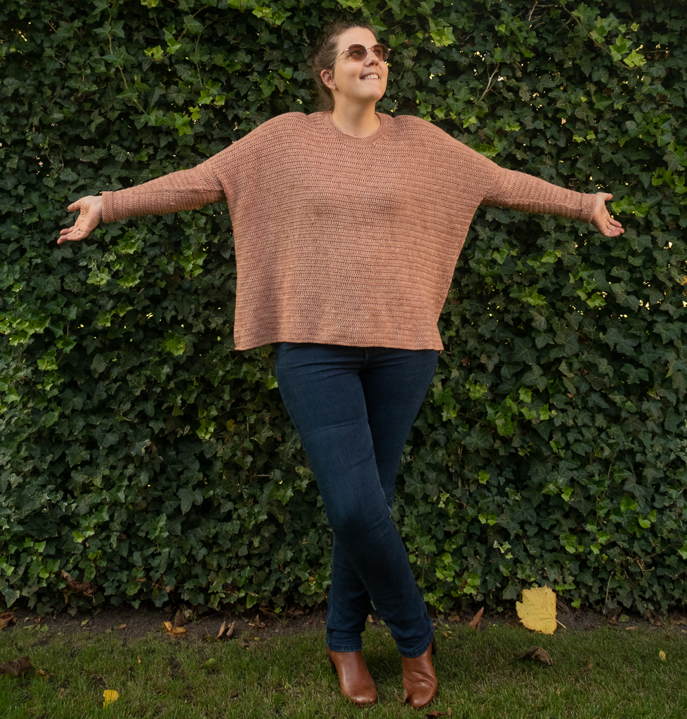 Woman standing up with her arms opened, showcasing a pinkish oversized sweater crochet pattern free, on a green natural background