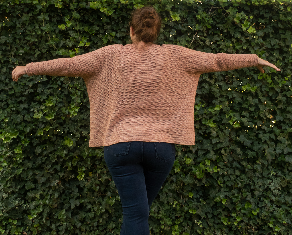 Woman standing up with arms wide opened to show the back panel of a pinkish peach oversized sweater crochet pattern free with blue jeans on a green natural background