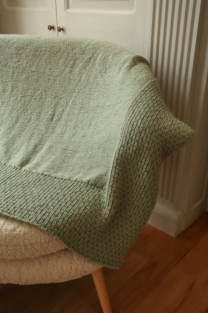 A green gender neutral baby blanket knitting pattern, thrown over a pale couch