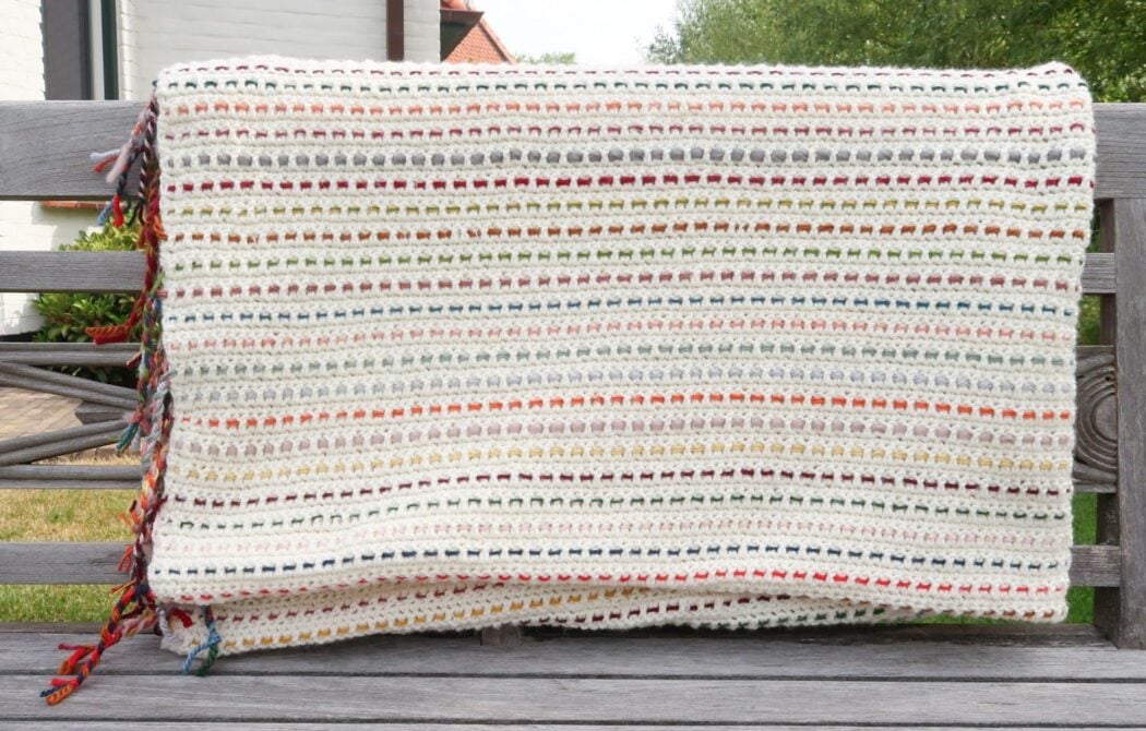 White stash buster crochet blanket with colorful woven threads draped on a bench