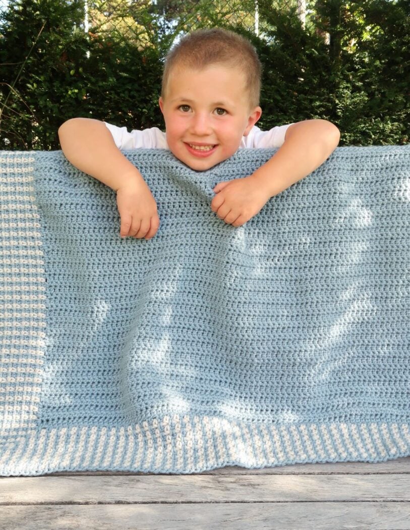 Moss stitch crochet baby blanket in blue and cream draped over a child's shoulders, from the moss stitch crochet blanket pattern